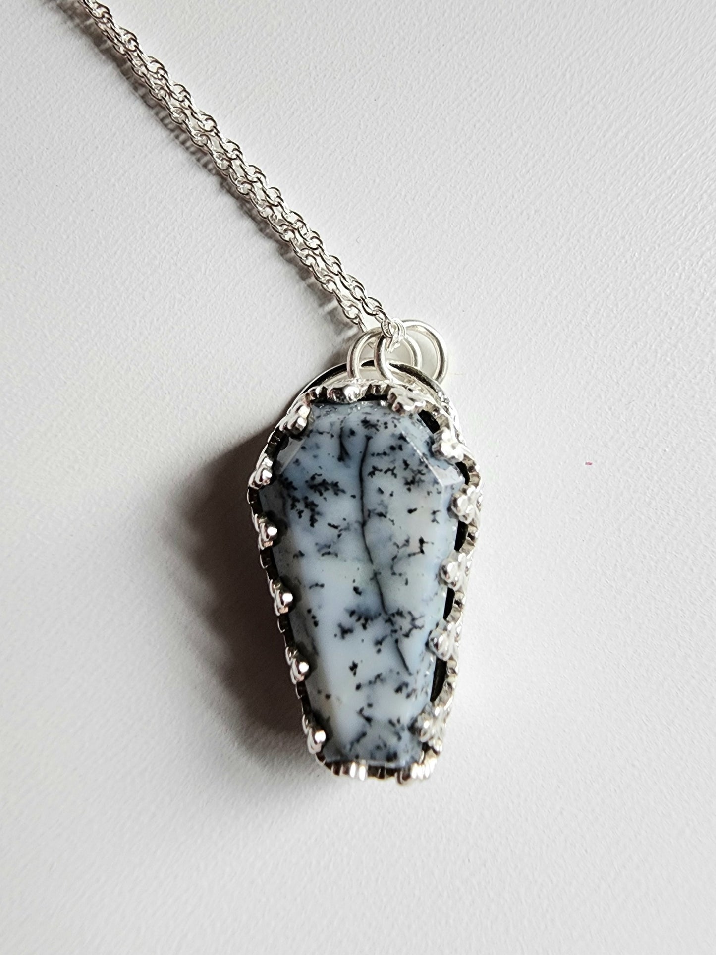 Dendritic agate coffin shaped stone mounted on a 18 inch sterling silver necklace by two jump rings. Stone is white with black variations surrounded by a pront castle style bezel wall that holds the stones in