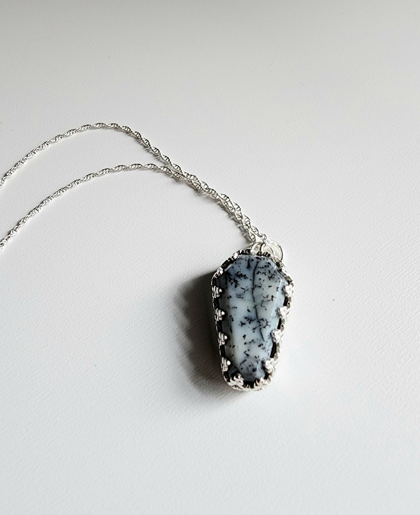Dendritic agate coffin shaped stone mounted on a 18 inch sterling silver necklace by two jump rings. Stone is white with black variations surrounded by a pront castle style bezel wall that holds the stones in