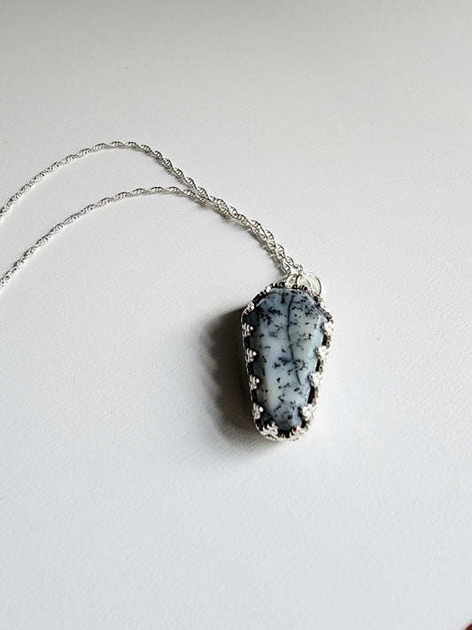 Dendritic agate coffin shaped stone mounted on a 18 inch sterling silver necklace by two jump rings. Stone is white with black variations surrounded by a pront castle style bezel wall that holds the stones in