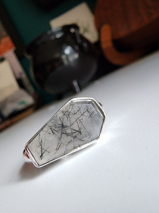 white stone with black flecks with metallic coffin stone shaped ring with thick band that meets the bezel on the left and right