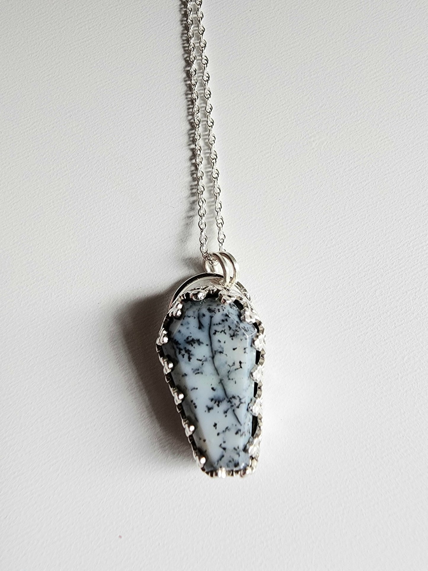 Dendritic agate coffin shaped stone mounted on a 18 inch sterling silver necklace by two jump rings. Stone is white with black variations surrounded by a pront castle style bezel wall that holds the stones in 