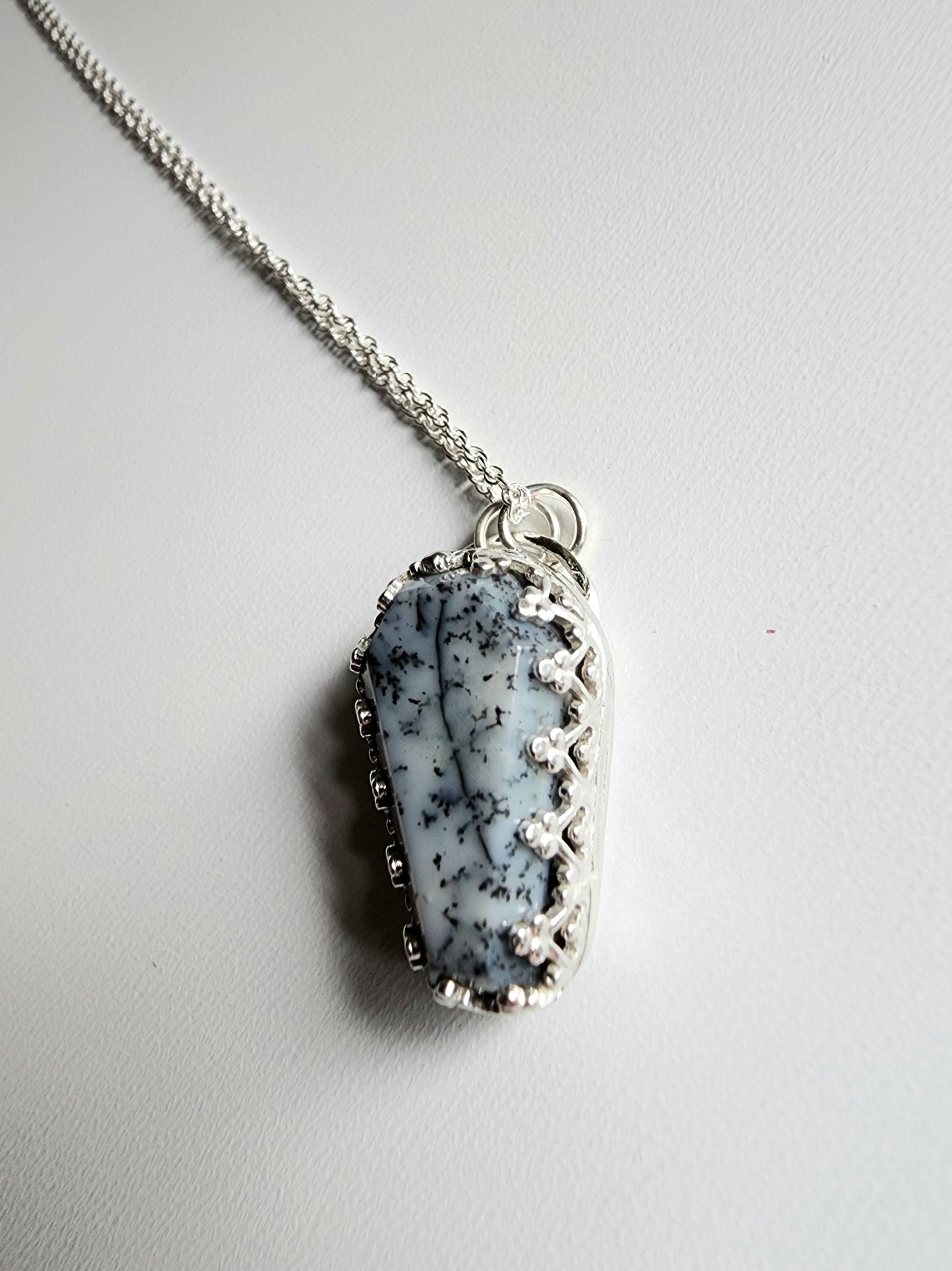 Dendritic agate coffin shaped stone mounted on a 18 inch sterling silver necklace by two jump rings. Stone is white with black variations surrounded by a pront castle style bezel wall that holds the stones in 