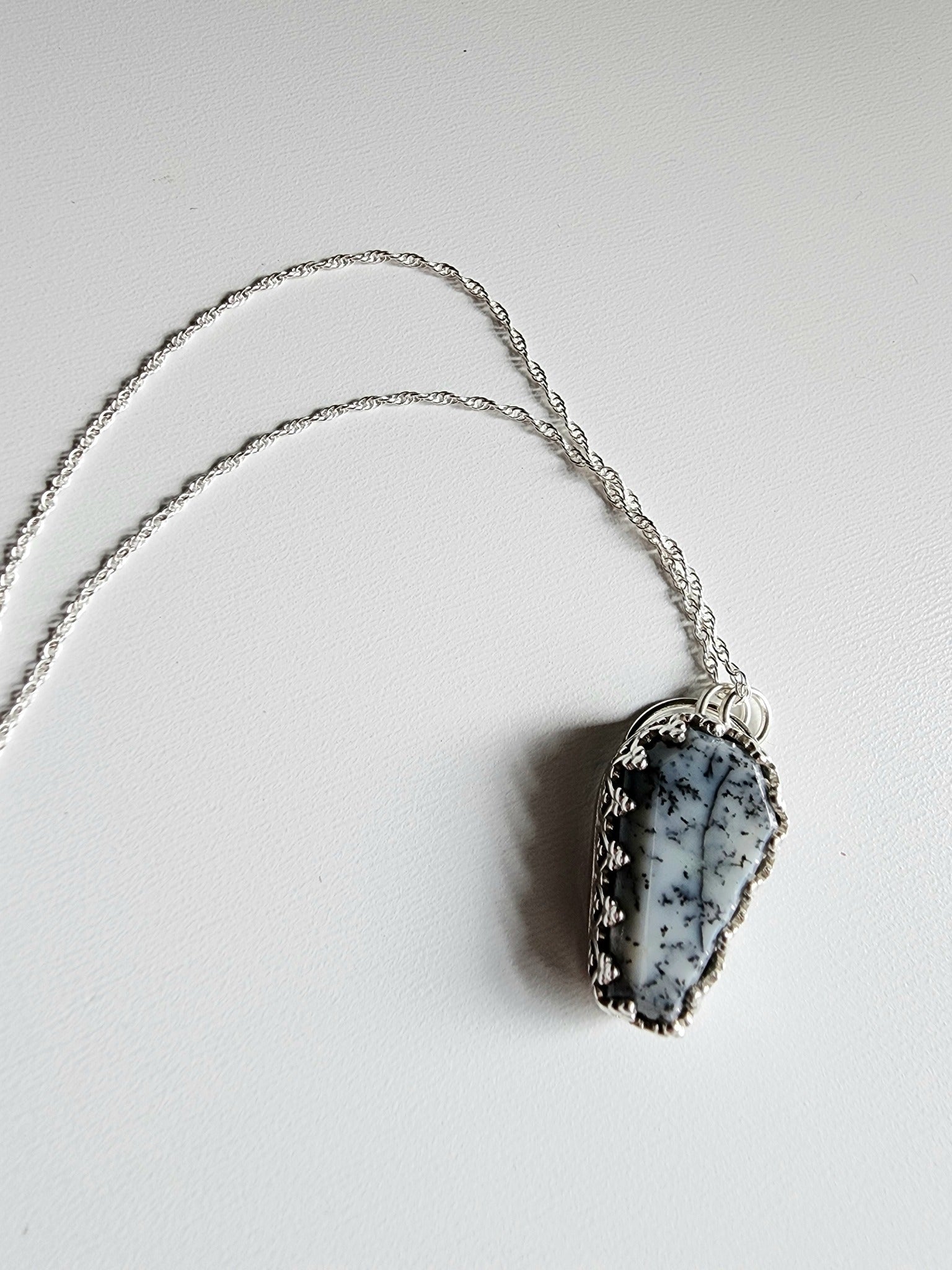 Dendritic agate coffin shaped stone mounted on a 18 inch sterling silver necklace by two jump rings. Stone is white with black variations surrounded by a pront castle style bezel wall that holds the stones in 