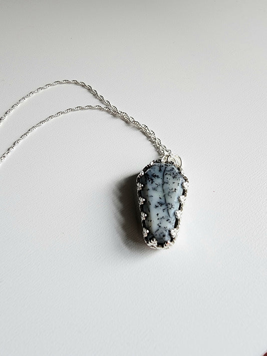 Dendritic agate coffin shaped stone mounted on a 18 inch sterling silver necklace by two jump rings. Stone is white with black variations surrounded by a pront castle style bezel wall that holds the stones in 