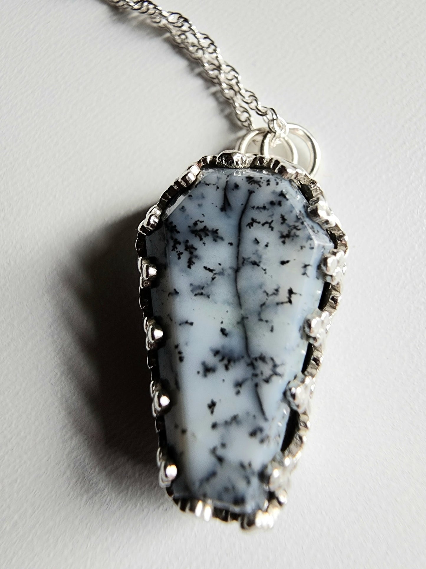 Dendritic agate coffin shaped stone mounted on a 18 inch sterling silver necklace by two jump rings. Stone is white with black variations surrounded by a pront castle style bezel wall that holds the stones in 