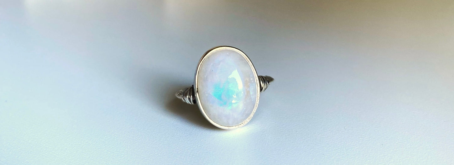 Large moonstone ring with scrollwork single band, left and right of the stone are small sterling silver wire wrapped accents. The moonstone itself has a gorgeous blue flash when the stone is met with direct sunlight. Surrounded by a simple bezel wall to protect the stone.