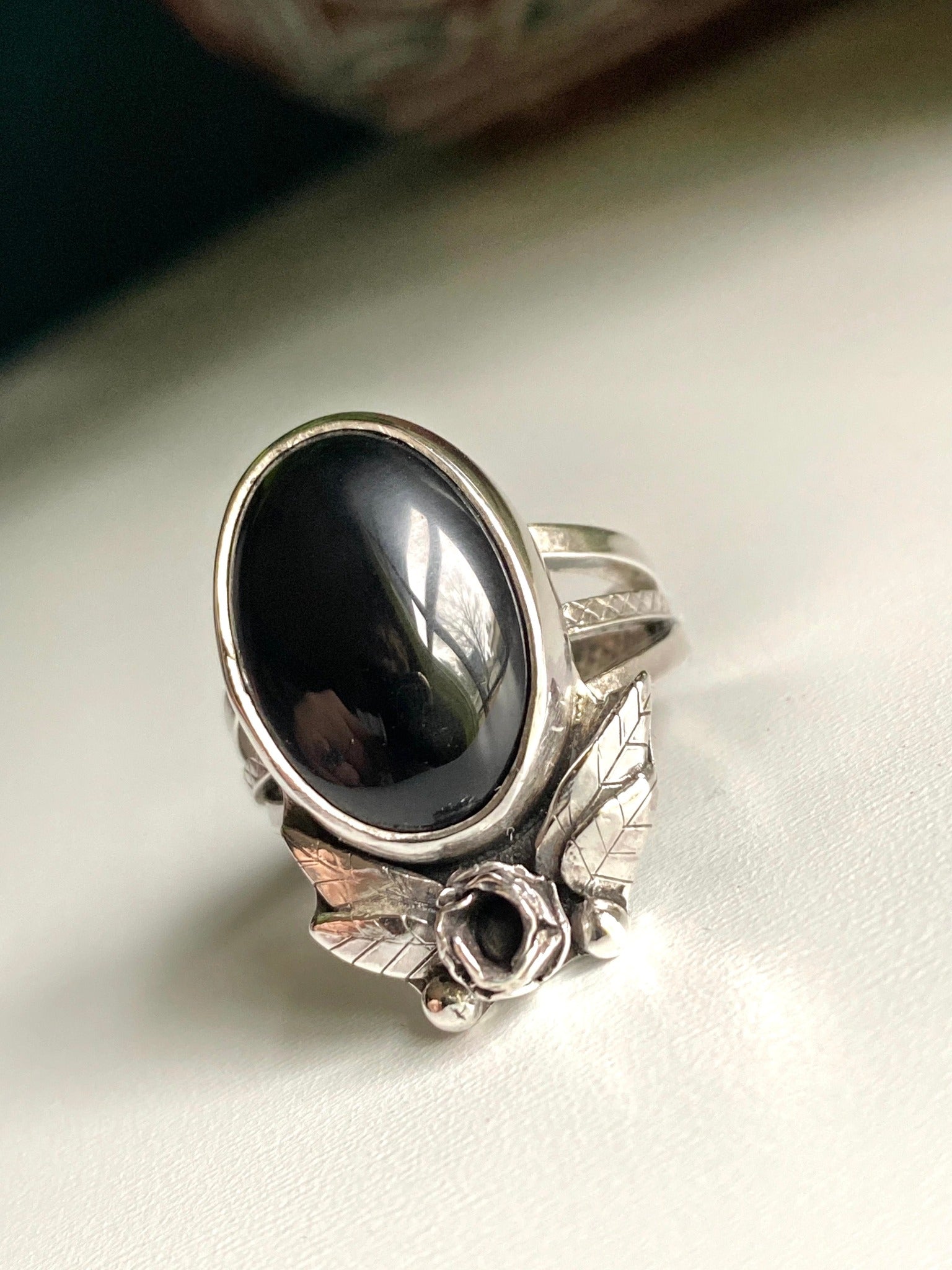 Onyx rose statement ring, Large onyx stone with rose below surrounded by 2 petals on each side, four total. hand carved leaves with two pieces of granulation at bottom. Ring is a triple strand shank two smooth square bands and one square middle band with tiny detail work looking like lattice.